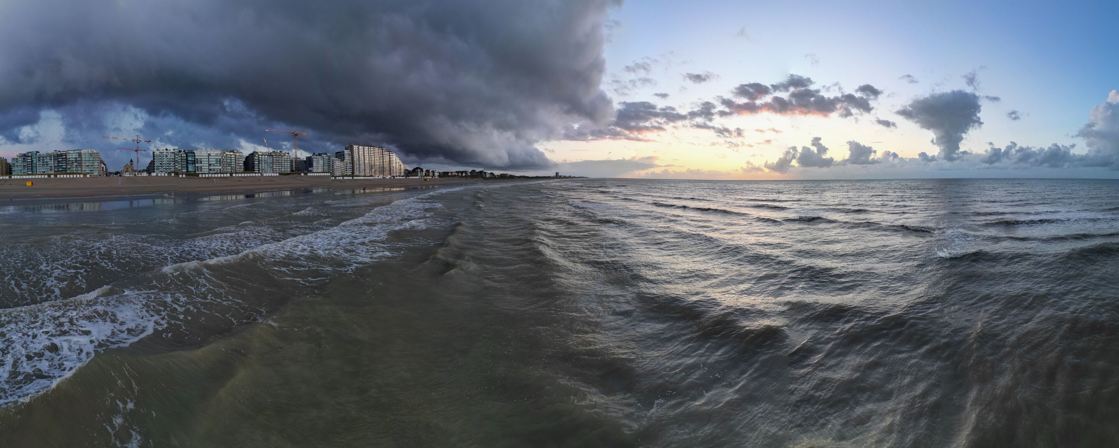 A beach with waves, a cloudy sky, and buildings in the background.