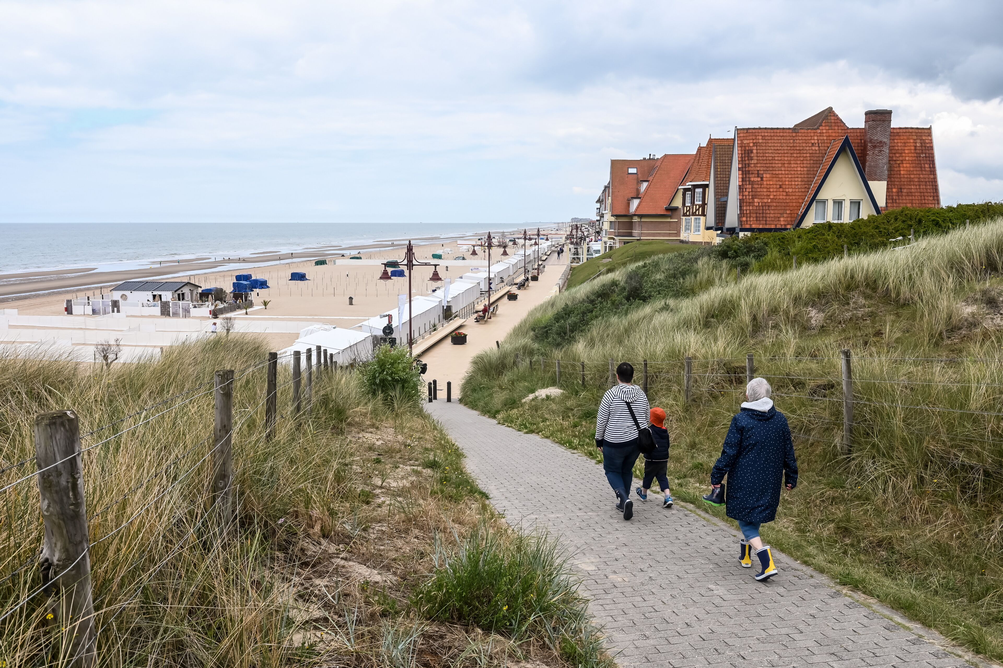 A coastal path leading to a beach with dunes, a wooden fence, and a building with a red roof.