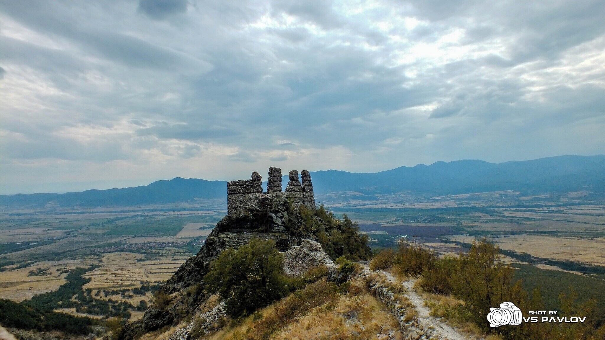 A stone structure on a hill with a valley and mountains in the background.