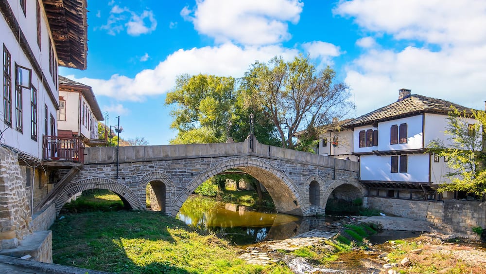 Eine Steinbrücke über einem Fluss, umgeben von traditioneller Architektur und viel Grün.