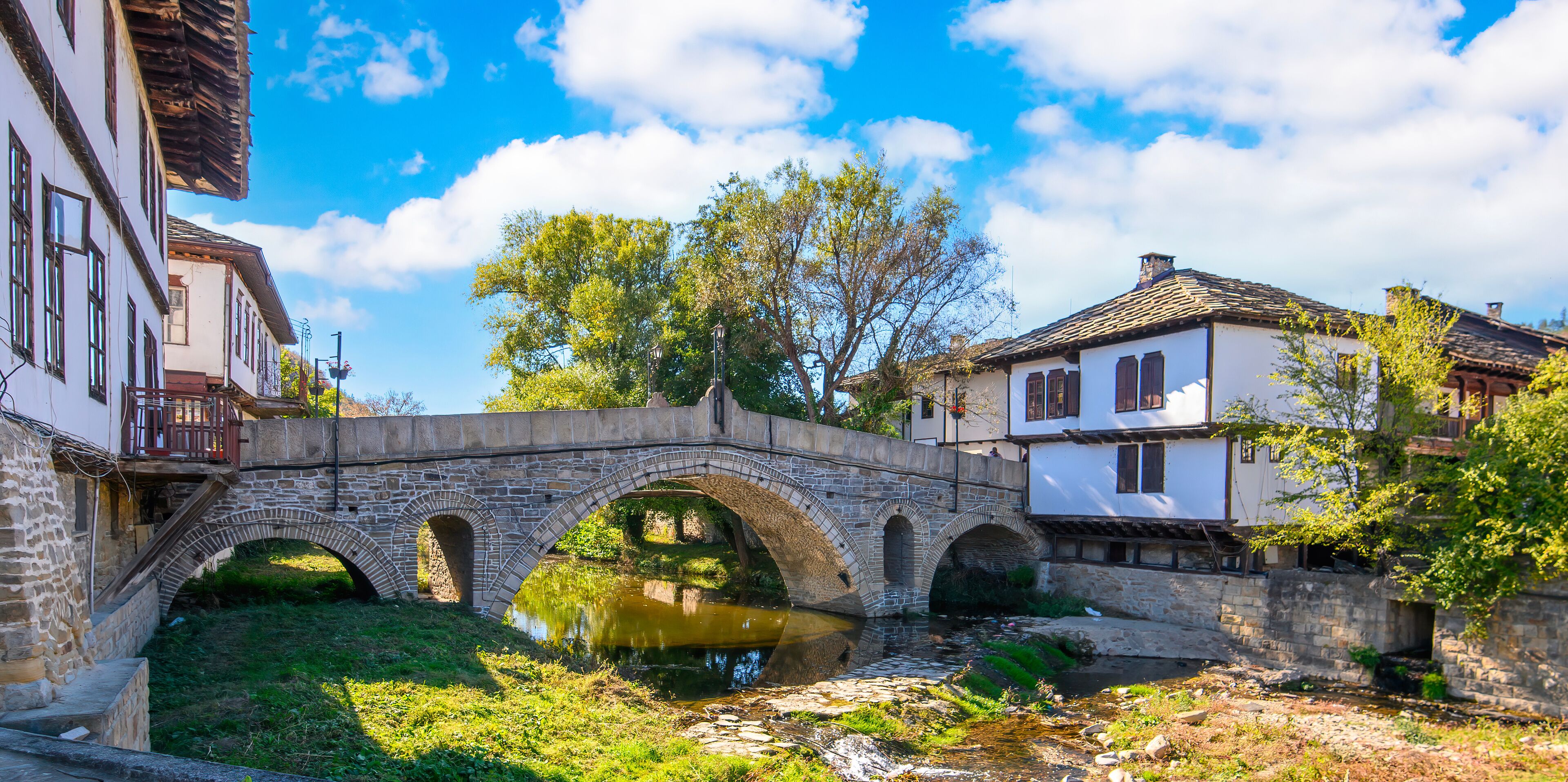 A stone bridge over a river, surrounded by traditional buildings and greenery.