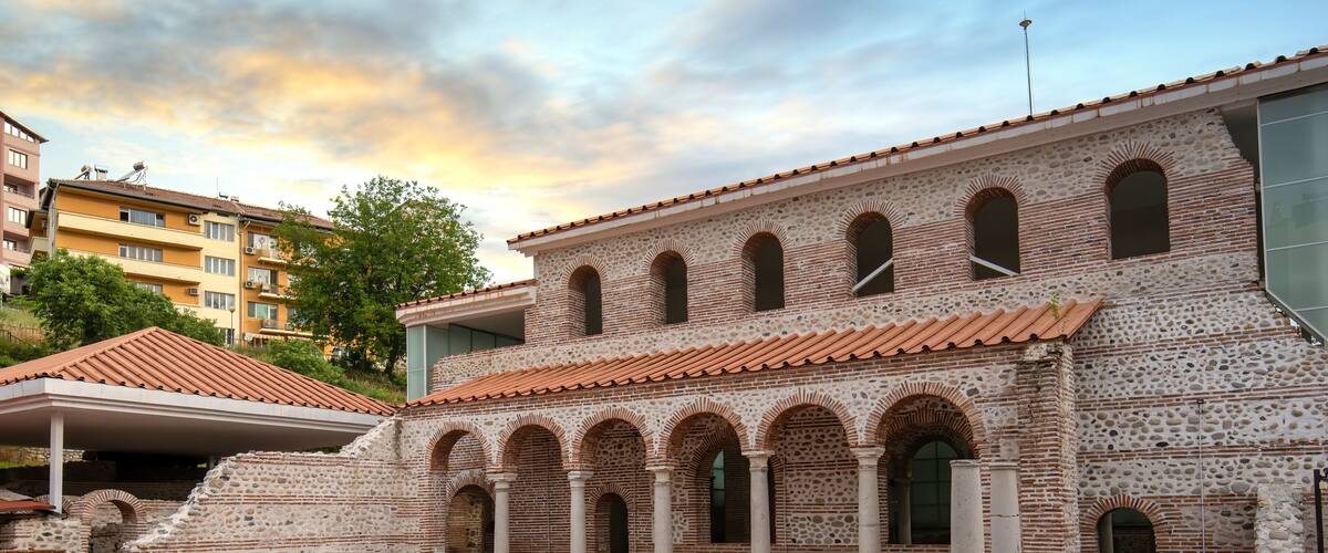 An ancient courtyard with stone columns and arches, surrounded by modern buildings.