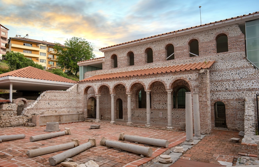An ancient courtyard with stone columns and arches, surrounded by modern buildings.