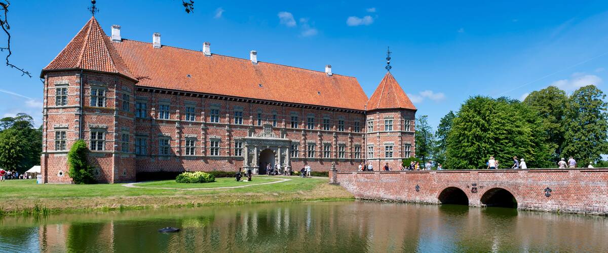 A large brick building with a red-tiled roof, a pond in front, and a bridge with three arches.