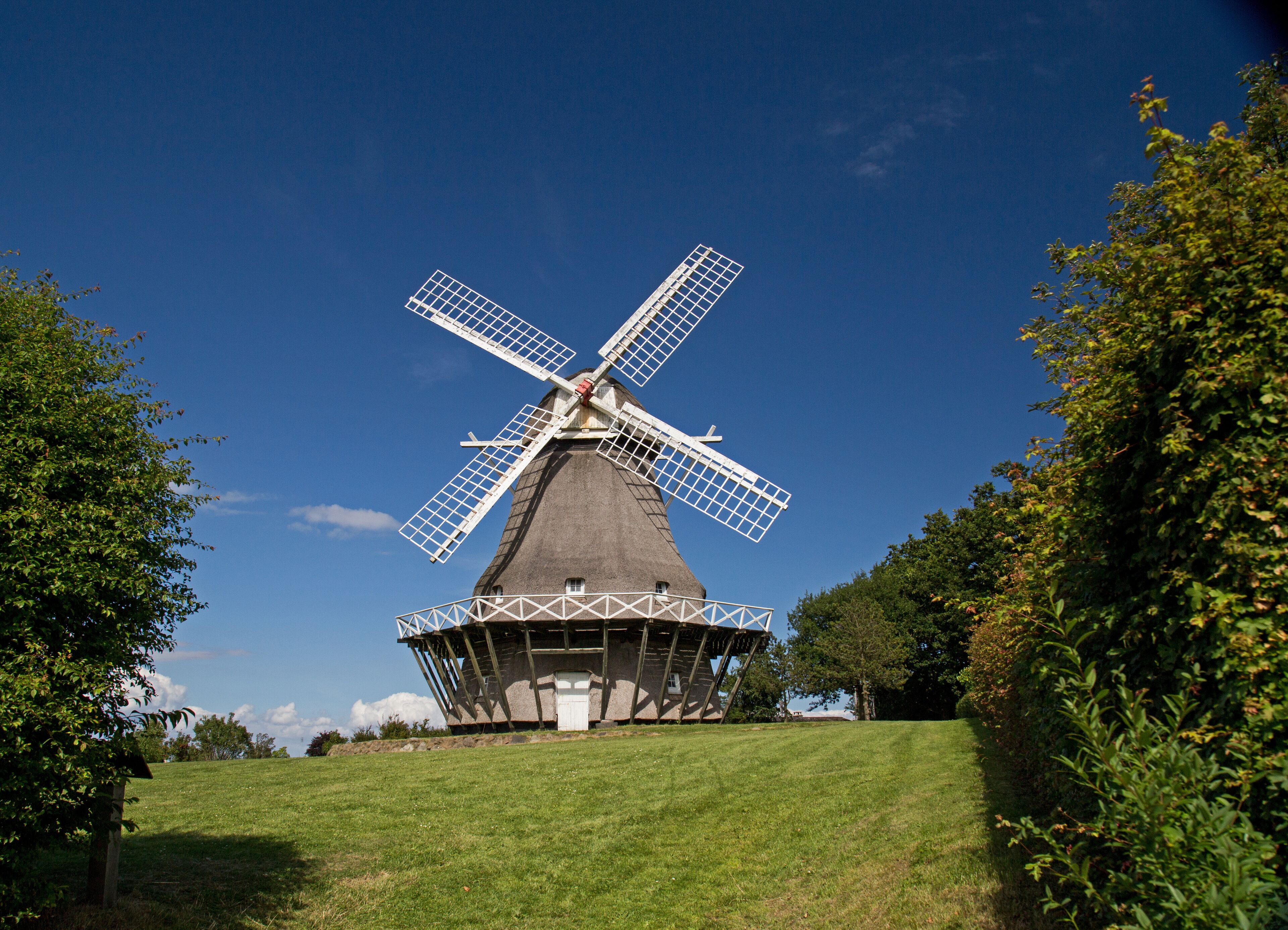 A traditional windmill with a large sail structure, surrounded by lush greenery and a clear blue sky.