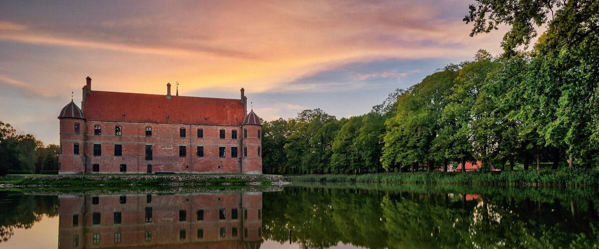 A large brick building with a red roof, reflecting in a calm body of water, surrounded by lush green trees.