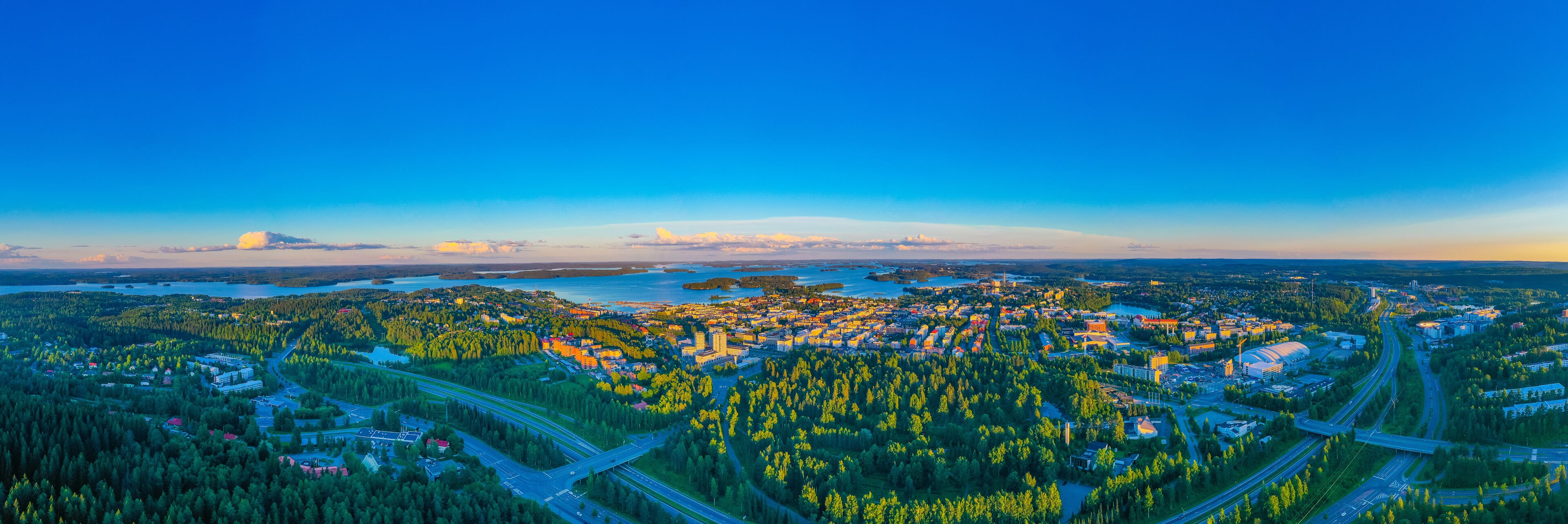 A coastal cityscape with dense forested hills, a river, and a large body of water.