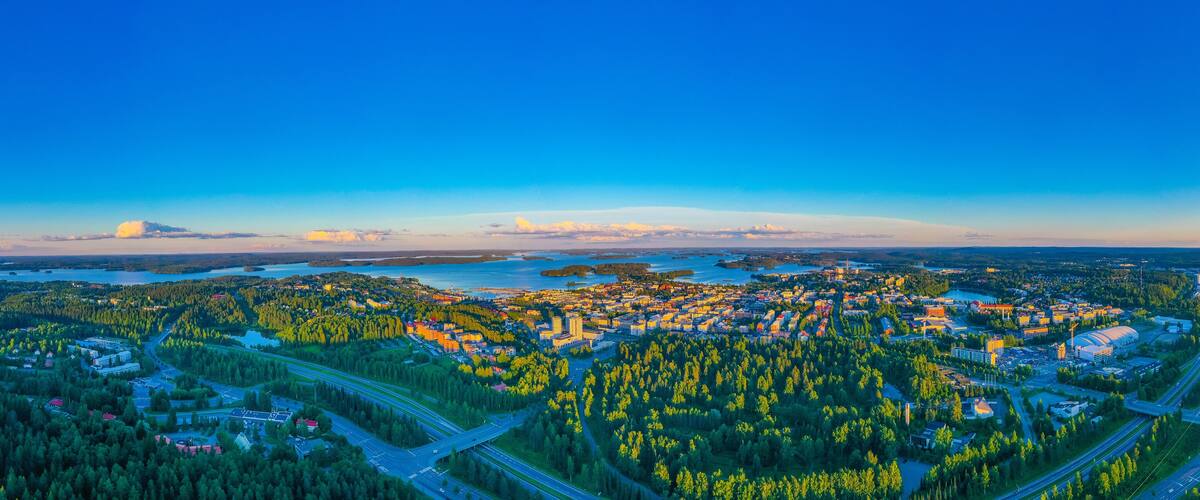 A coastal cityscape with dense forested hills, a river, and a large body of water.