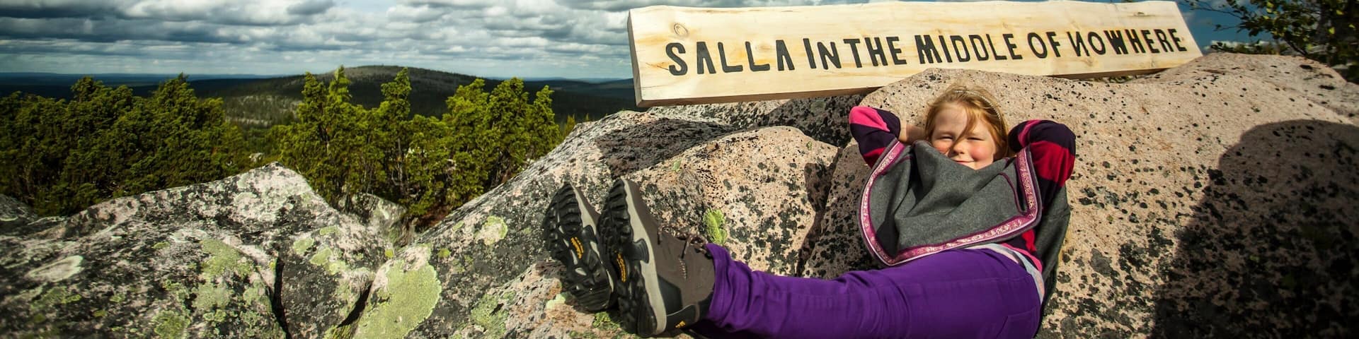 A person sitting on a rock with a sign that reads "SALLA IN THE MIDDLE OF NOWHERE".