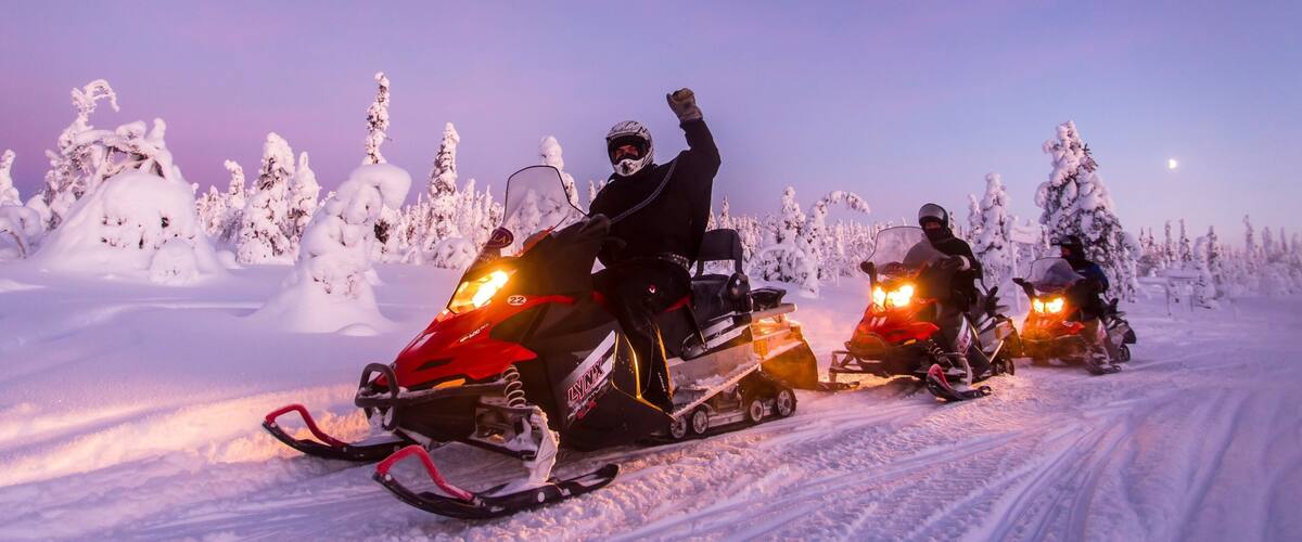 Three snowmobiles on a snowy path with snow-covered trees and a purple sky.
