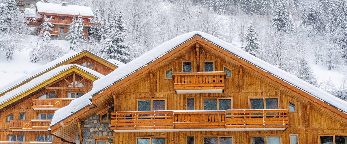 A wooden chalet with a stone foundation, surrounded by snow-covered trees and a forested mountain backdrop.