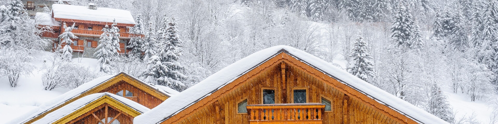 A wooden chalet with a stone foundation, surrounded by snow-covered trees and a forested mountain backdrop.