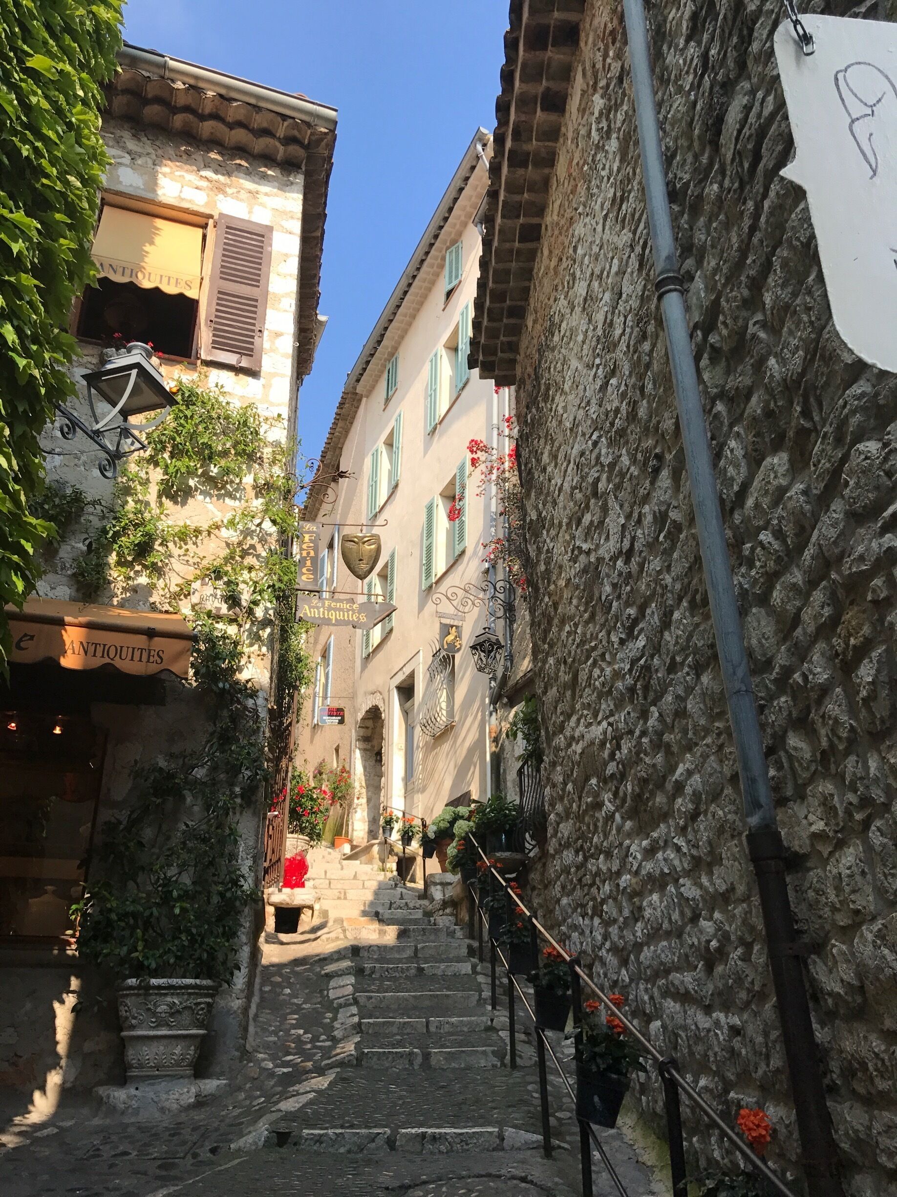 A narrow cobblestone street flanked by historic buildings with shutters and a sign for 'Antiquités'.