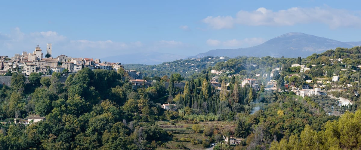 A town with historic buildings and a prominent tower, surrounded by lush green hills and a mountain in the background.