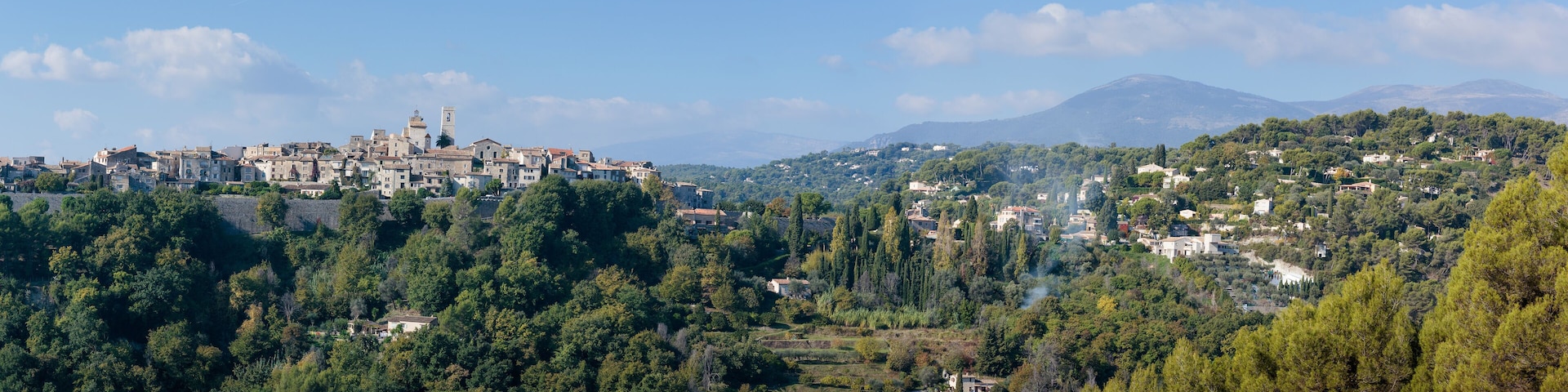 A town with historic buildings and a prominent tower, surrounded by lush green hills and a mountain in the background.