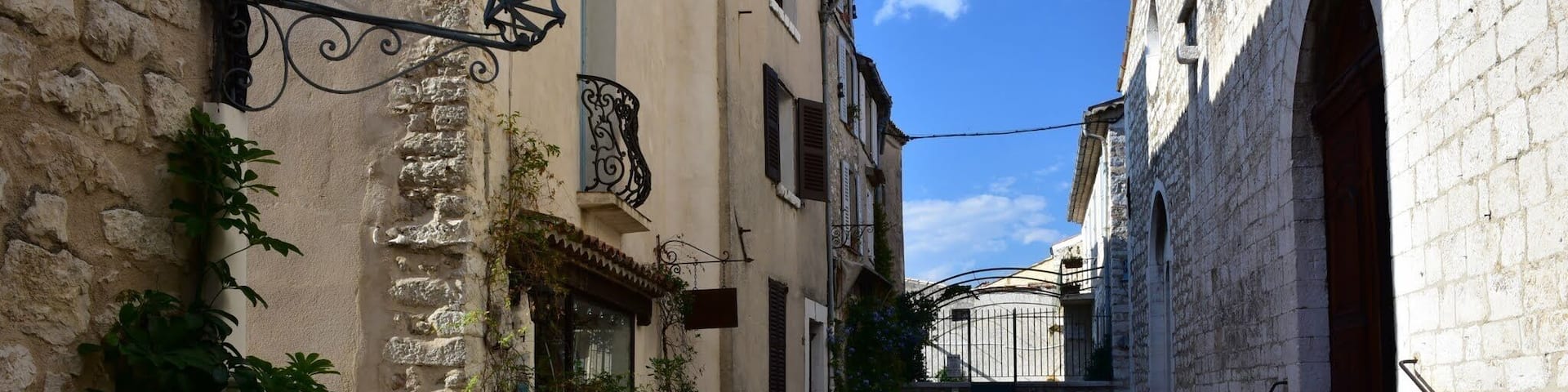 A narrow stone pathway flanked by historic buildings with arched doorways and a staircase leading up to a gate.
