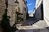 A narrow stone pathway flanked by historic buildings with arched doorways and a staircase leading up to a gate.