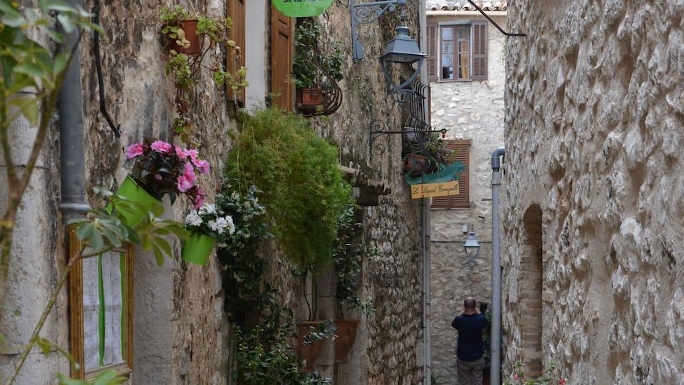 Une ruelle étroite pavée, avec des chaises et des tables en métal vert, des plantes en pot et une enseigne indiquant un café-crêperie.