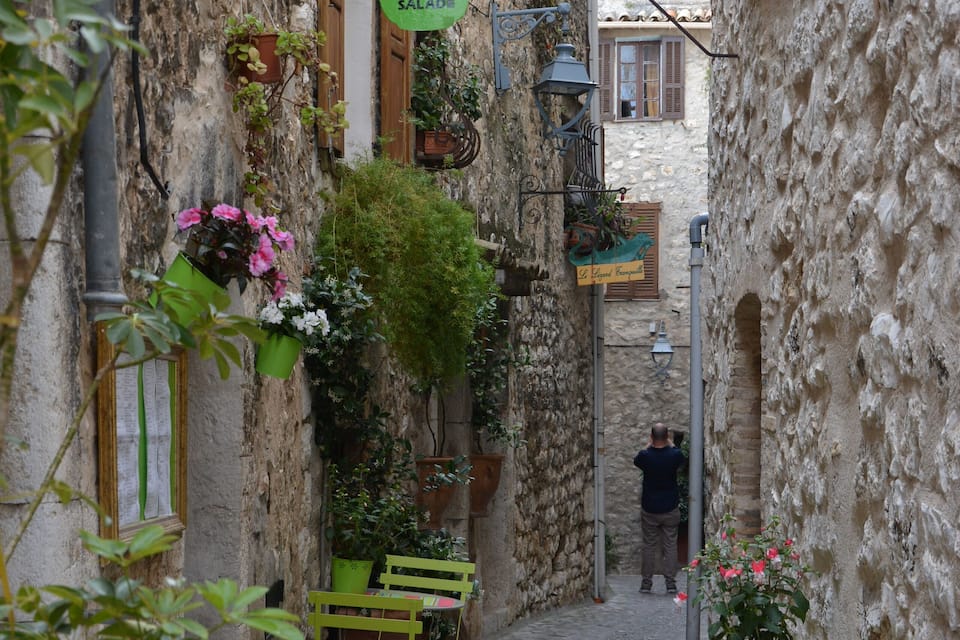 Une ruelle étroite pavée, avec des chaises et des tables en métal vert, des plantes en pot et une enseigne indiquant un café-crêperie.