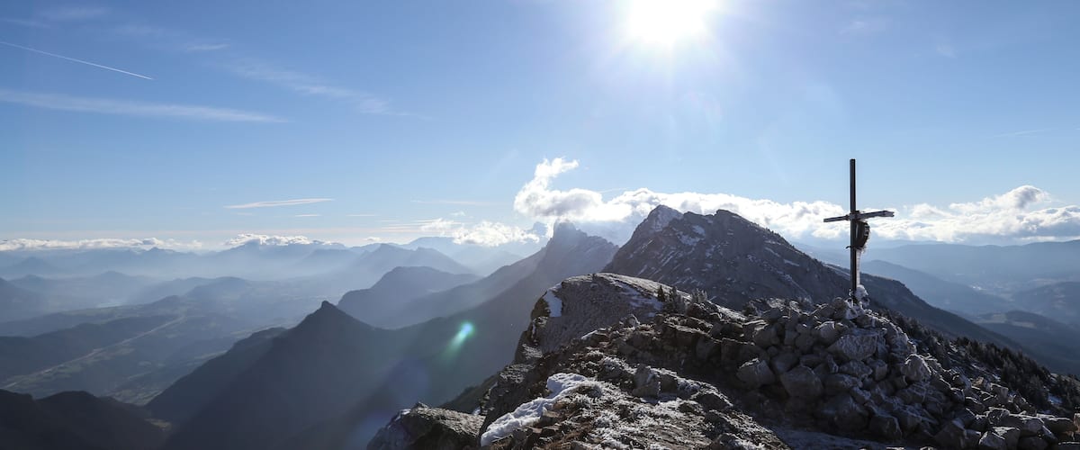 A mountain peak with a cross, snow-covered rocks, and a clear sky with the sun shining.