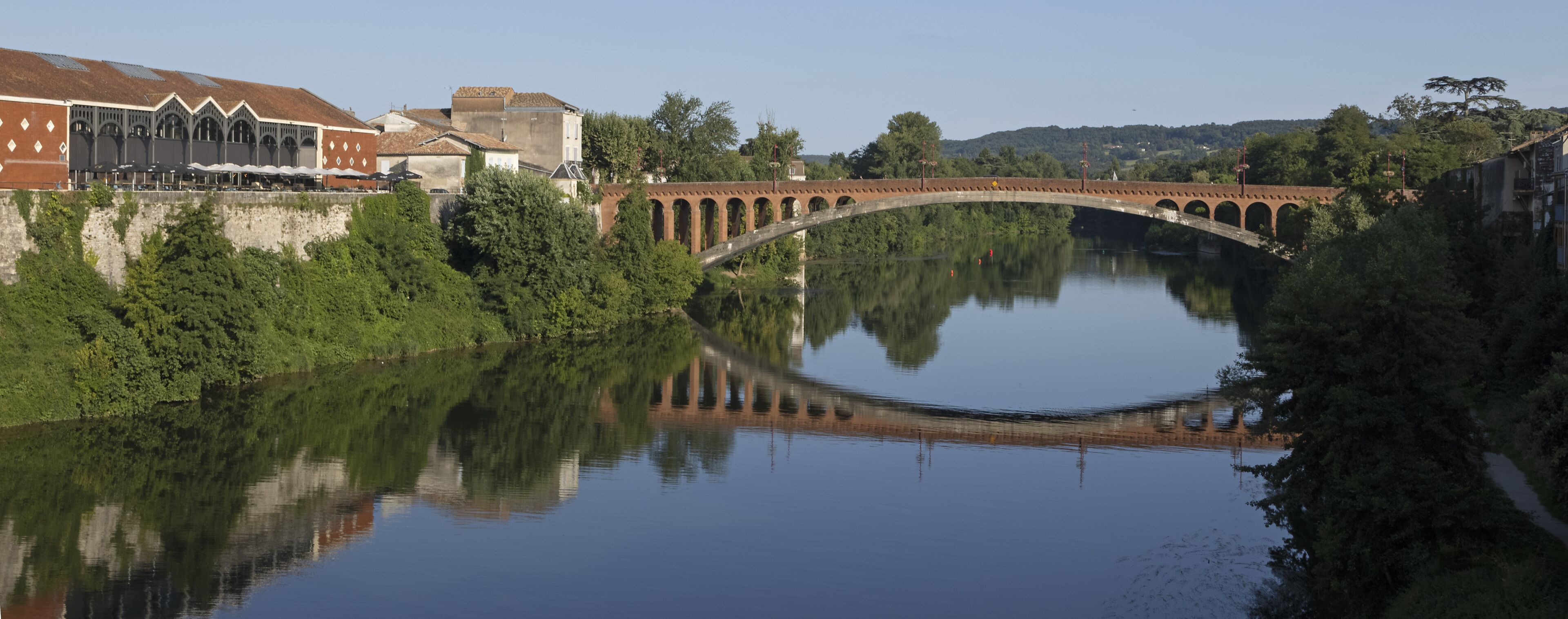 A stone bridge over a calm river, surrounded by lush greenery and buildings.