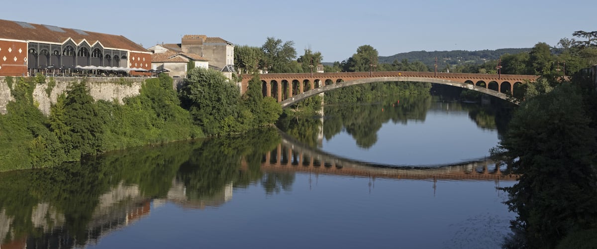 A stone bridge over a calm river, surrounded by lush greenery and buildings.