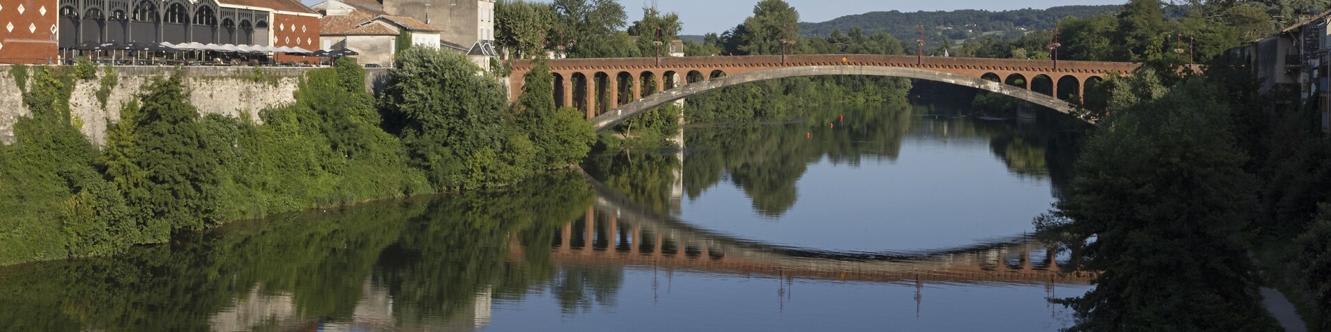 A stone bridge over a calm river, surrounded by lush greenery and buildings.