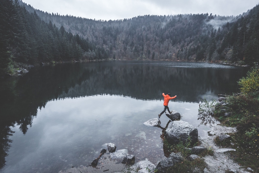 Une personne portant une veste orange marche sur des rochers au bord d’un lac paisible, entouré d’une forêt dense.