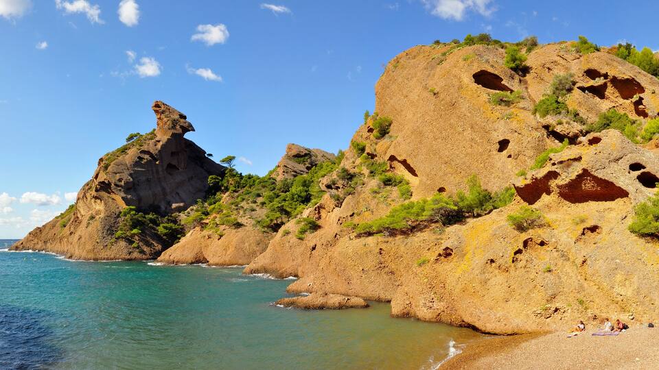 A coastal landscape with rocky cliffs, clear blue sea, and a few people on the beach.
