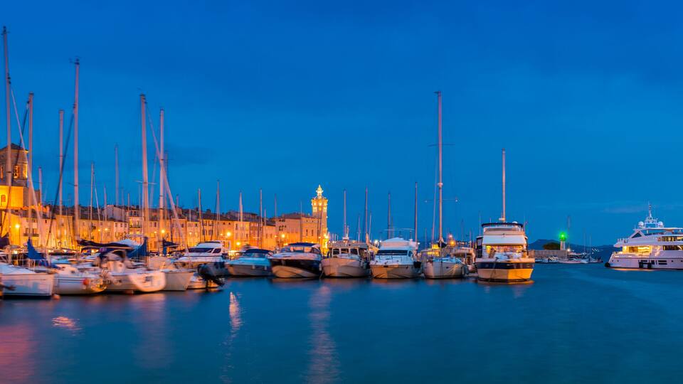 A marina at dusk with numerous boats docked, a prominent building with a tower, and a clear blue sky.