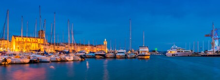 Un port de plaisance au crépuscule, avec de nombreux bateaux amarrés, un bâtiment imposant doté d’une tour, et un ciel bleu dégagé.