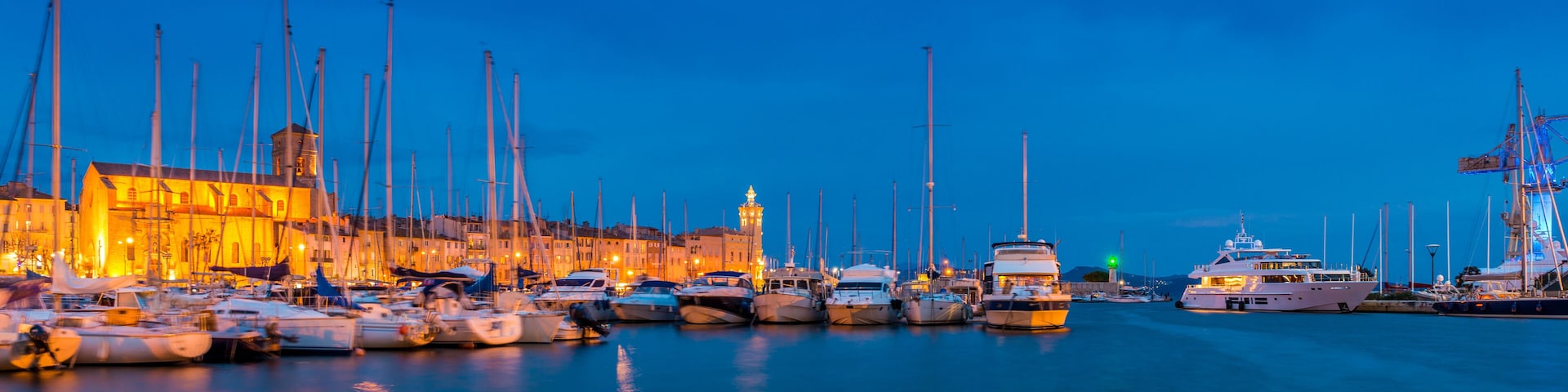 A marina at dusk with numerous boats docked, a prominent building with a tower, and a clear blue sky.