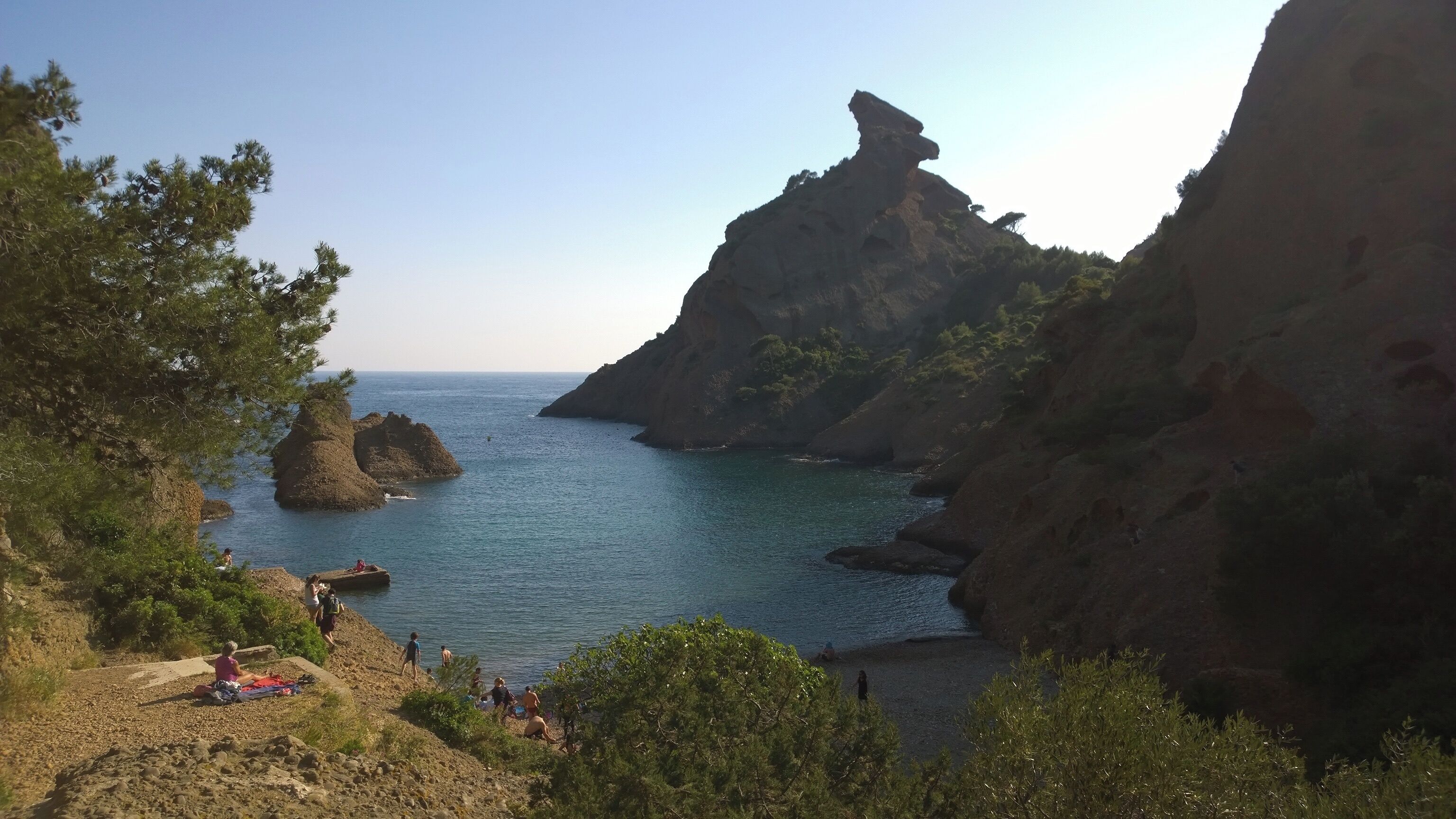 A coastal landscape with rocky cliffs, clear blue sea, and people enjoying the beach.