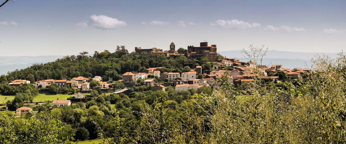 A village with red-roofed houses nestled in a valley, surrounded by greenery and a prominent castle on a hill.