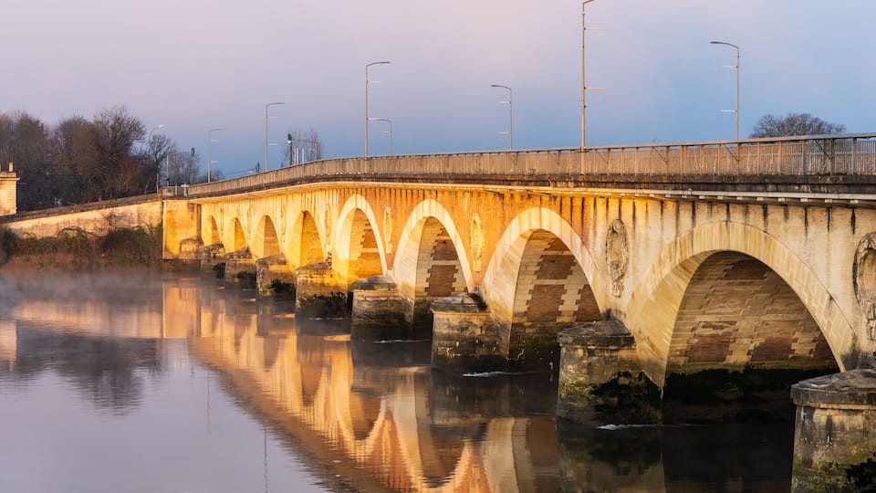 A stone bridge with multiple arches over a calm river, with street lamps and a clear sky in the background.