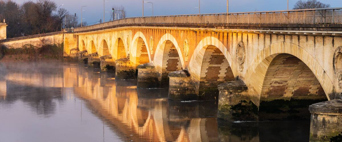 A stone bridge with multiple arches over a calm river, with street lamps and a clear sky in the background.