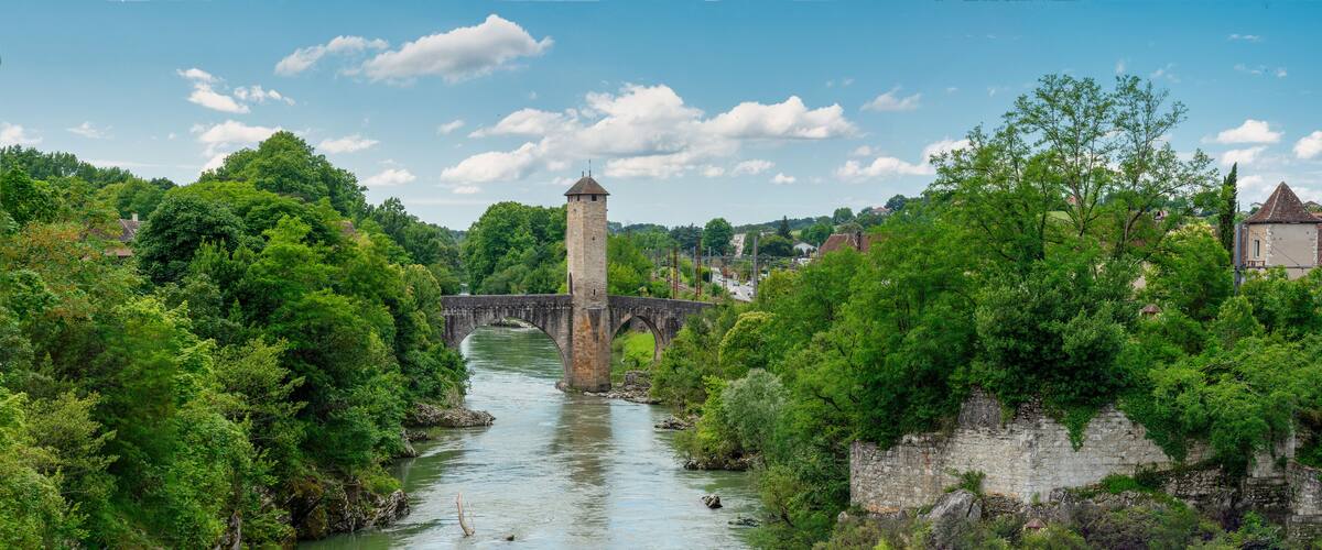 A stone bridge over a river, surrounded by lush greenery and a tower.