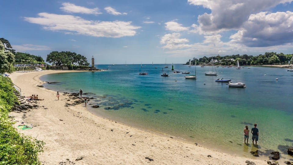 A beach with clear turquoise water, boats, and a lighthouse.