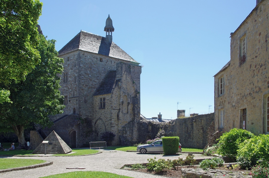 A historic stone building with a bell tower, surrounded by greenery and a parked car.