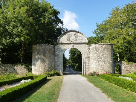 Un arc en pierre orné de blasons, entouré de jardins et d’arbres aux formes soignées.
