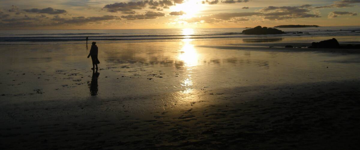 Een strand bij zonsondergang, met iemand die op het natte zand staat, de zon die over de horizon zakt en verre rotsen.