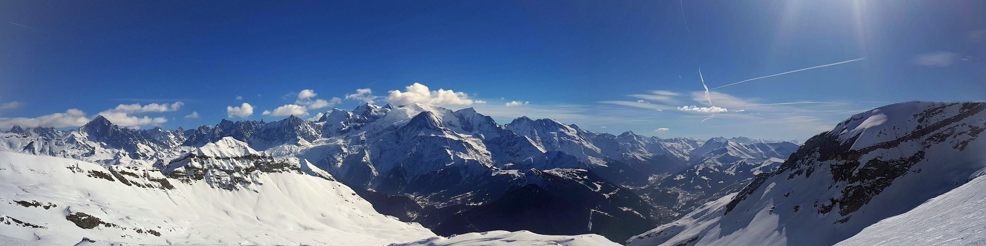A snow-covered mountain range with a clear blue sky and a visible contrail.