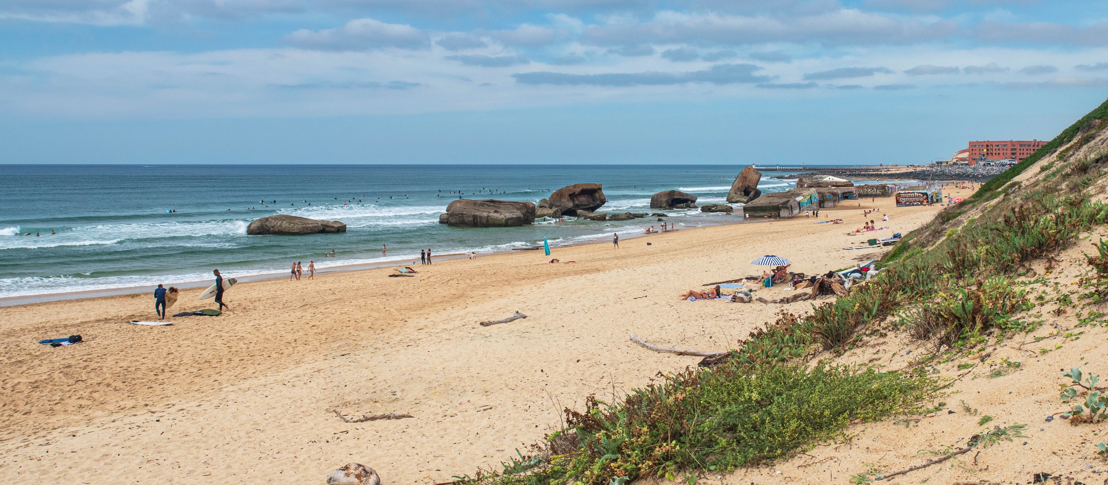 A sandy beach with large rocks, a clear sky, and a distant building.