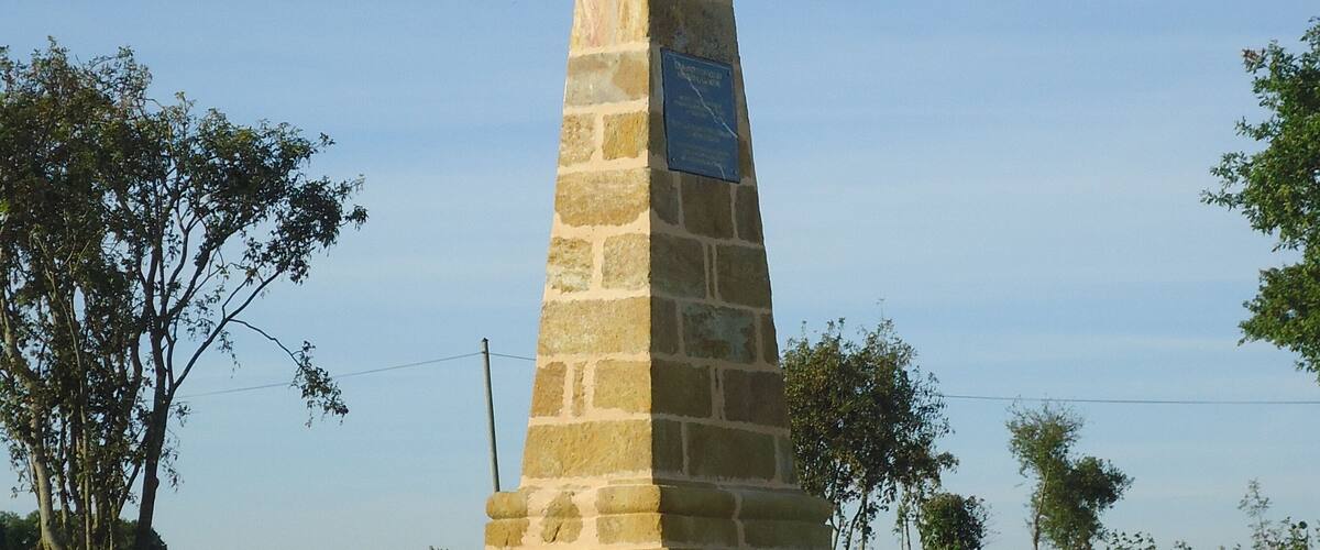 A stone monument with a plaque, surrounded by greenery and fields.