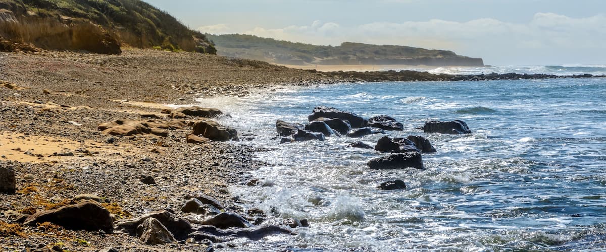 A rocky beach with waves crashing onto the shore, a distant coastline, and a hilly landscape under a partly cloudy sky.