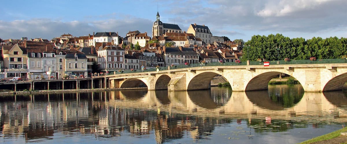 A stone bridge over a calm river with a town in the background.