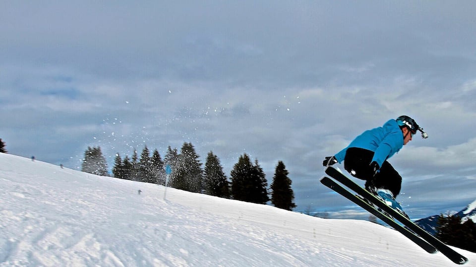 A skier in mid-air performing a jump on a snowy slope with trees and mountains in the background.