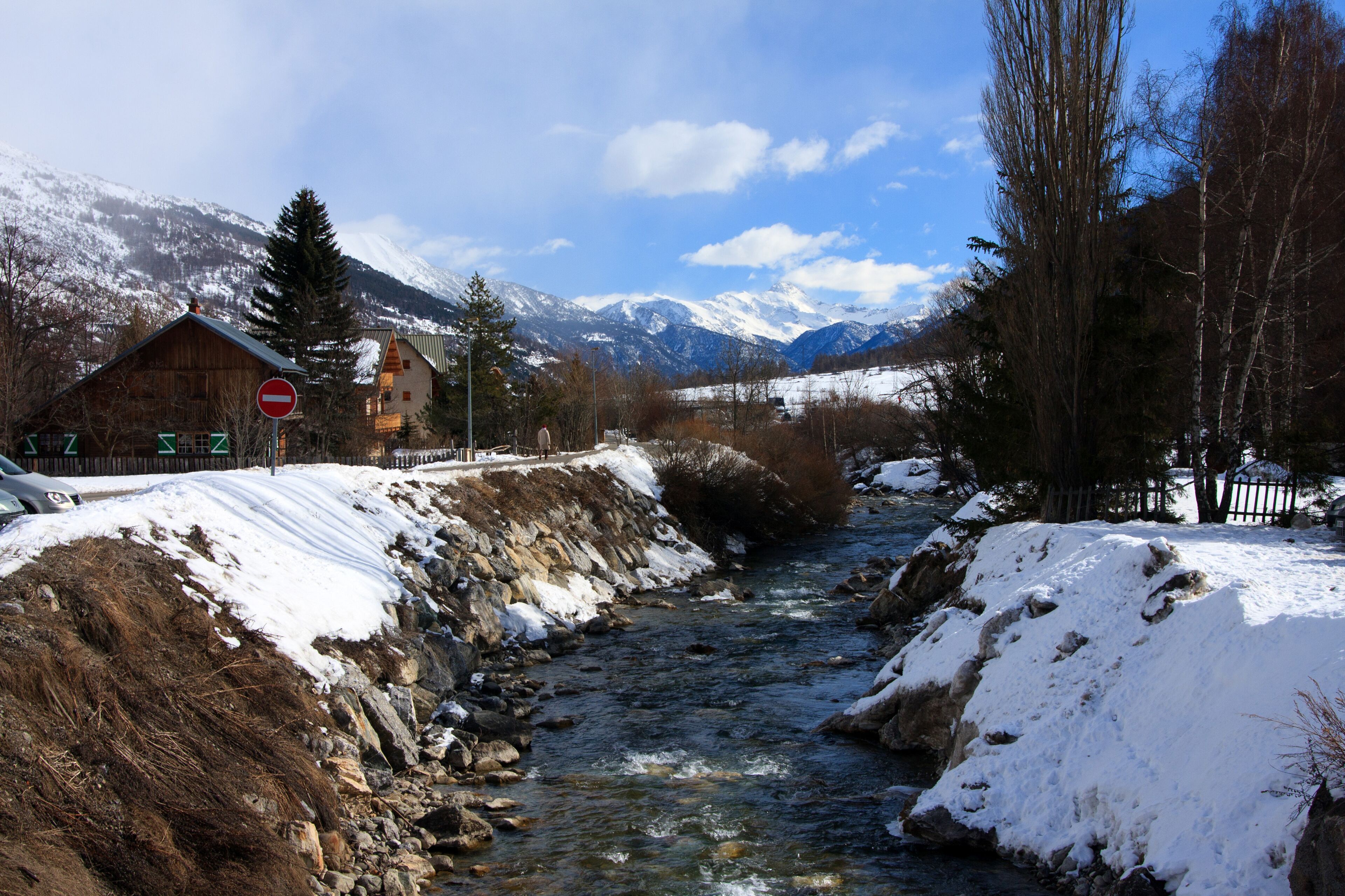 A snow-covered river with mountains in the background and a wooden house on the left.