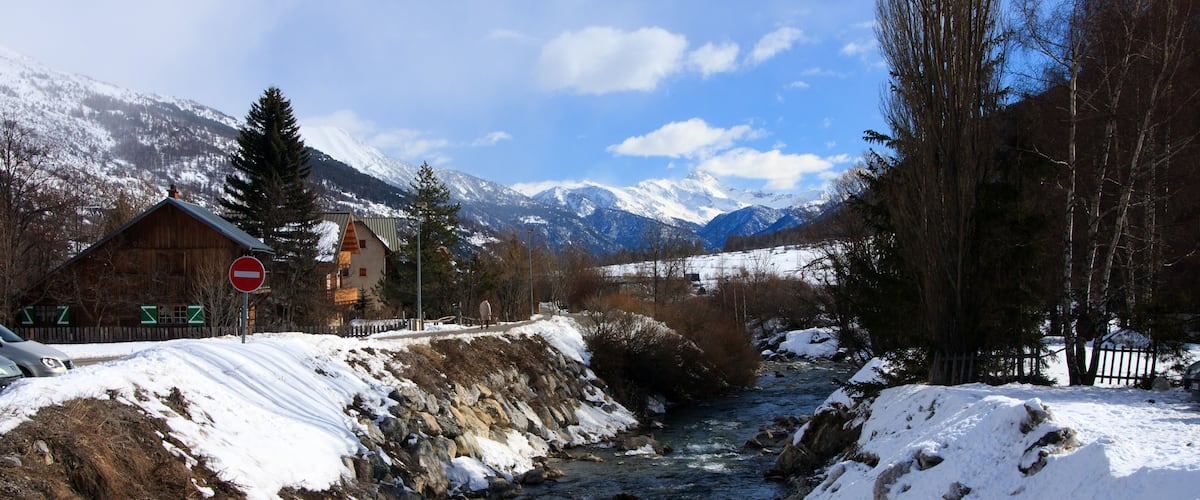 A snow-covered river with mountains in the background and a wooden house on the left.