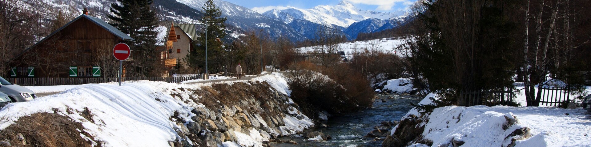 A snow-covered river with mountains in the background and a wooden house on the left.
